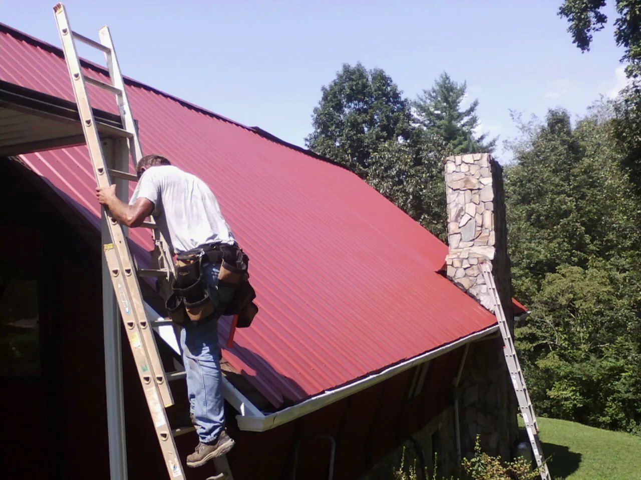 Roofer on a ladder beside a red metal roof and stone chimney — Soffit & Fascia Repair by Roofing Brothers in San Bruno